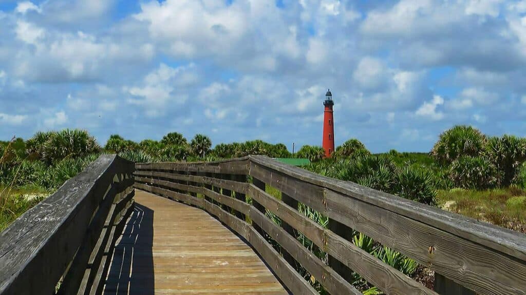 Boardwalk leading to red Ponce Inlet Lighthouse under a partly cloudy sky.