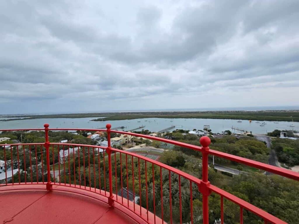 View from St Augustine Lighthouse, red observation deck overlooking water, boats, and greenery under a cloudy sky.