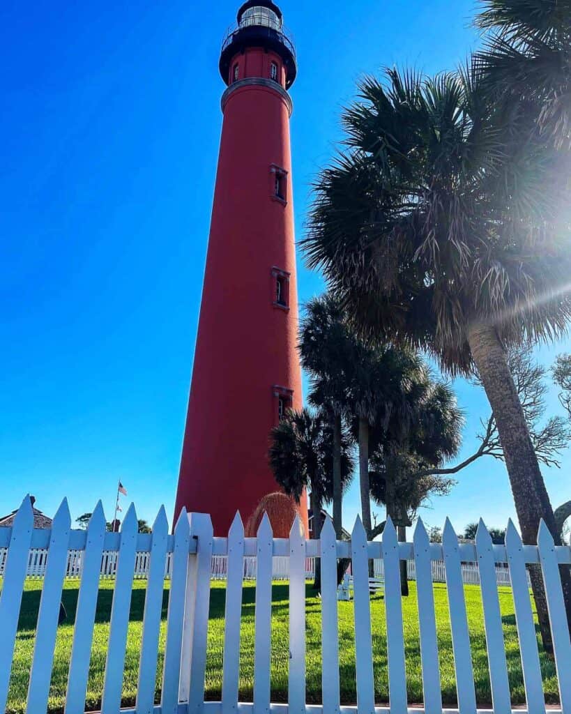 Ponce Inlet red lighthouse with a black top, surrounded by palm trees, behind a white picket fence under a blue sky.
