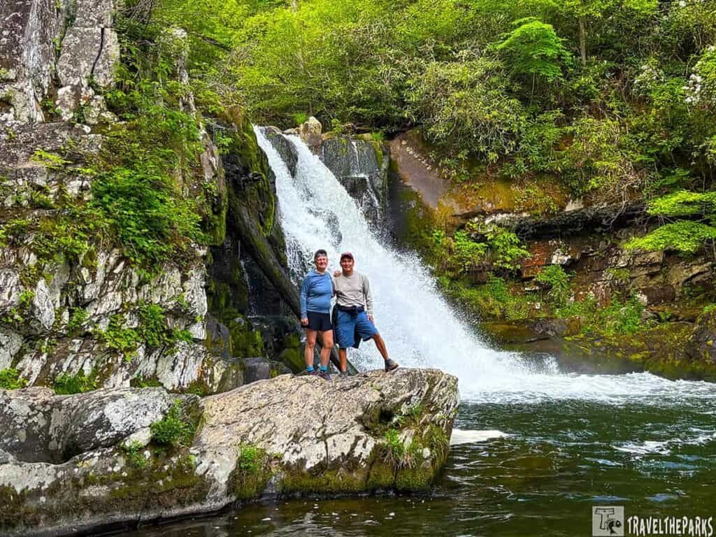 Two people standing on a rock in front of Abrams Falls, surrounded by greenery.