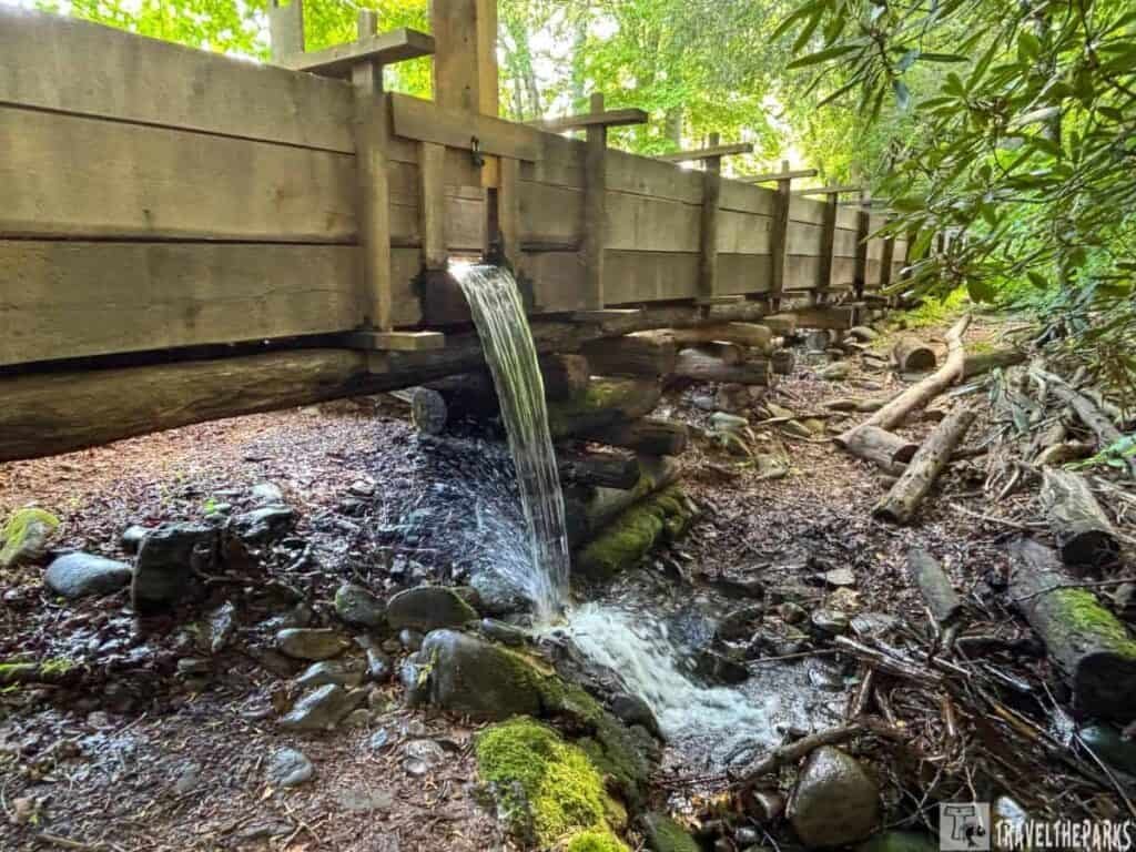Wooden flume with water flowing into a rocky streambed in a wooded area.

