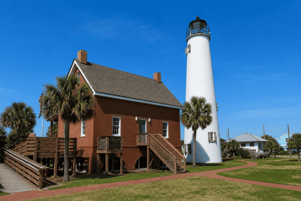 Cape St. George Lighthouse with adjacent red brick house, palm trees, and pathways under a clear blue sky.