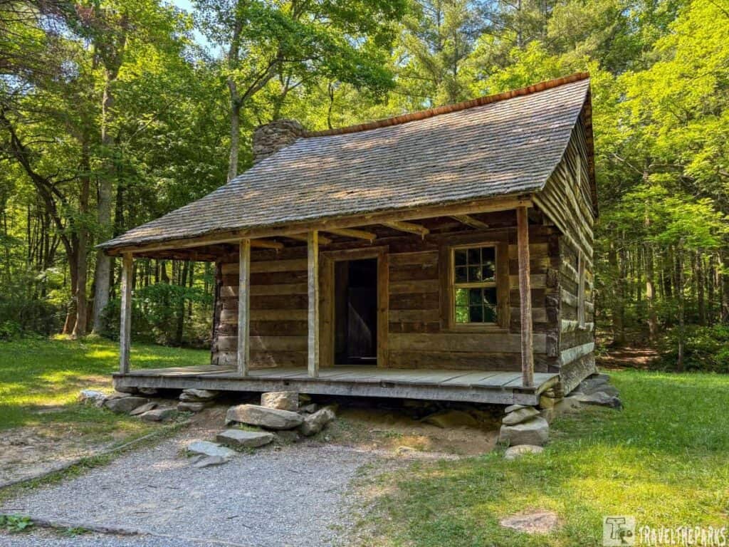 Rustic log cabin on the Cades Cove Loop Road in a wooded area with a stone chimney and front porch.