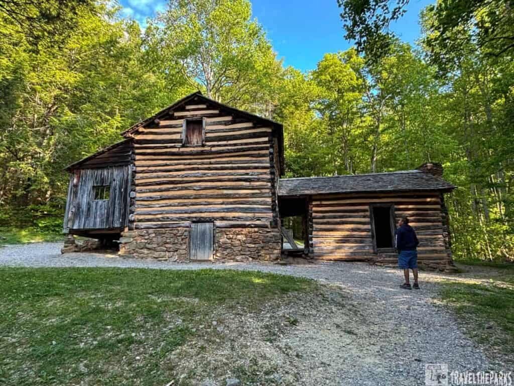 A historic log cabin in Cades Cove Elijah Oliver Place surrounded by greenery, with a person standing nearby. 