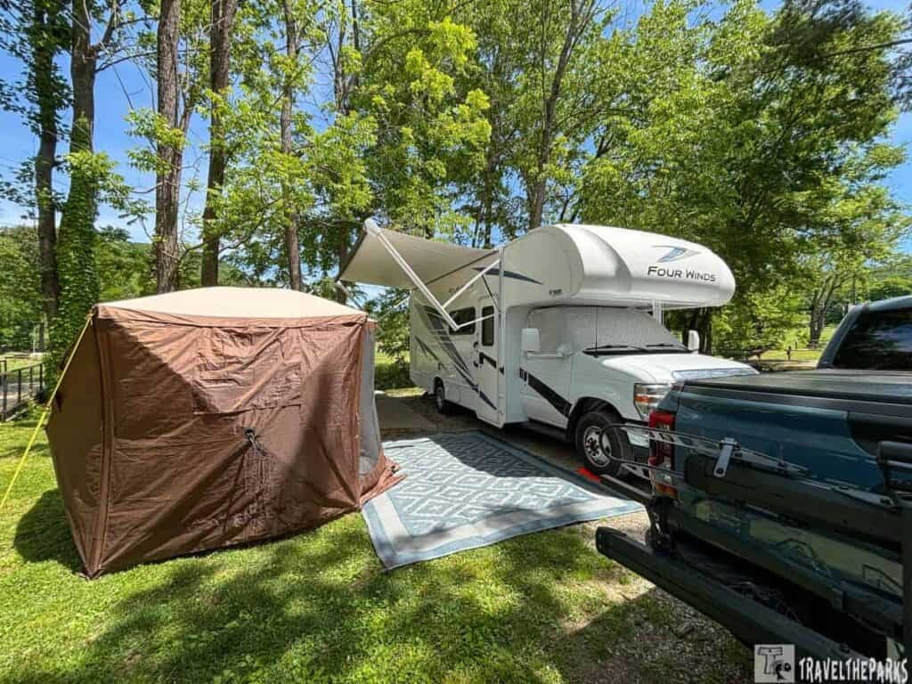 "RV and canopy tent in wooded campground with clear blue sky."

