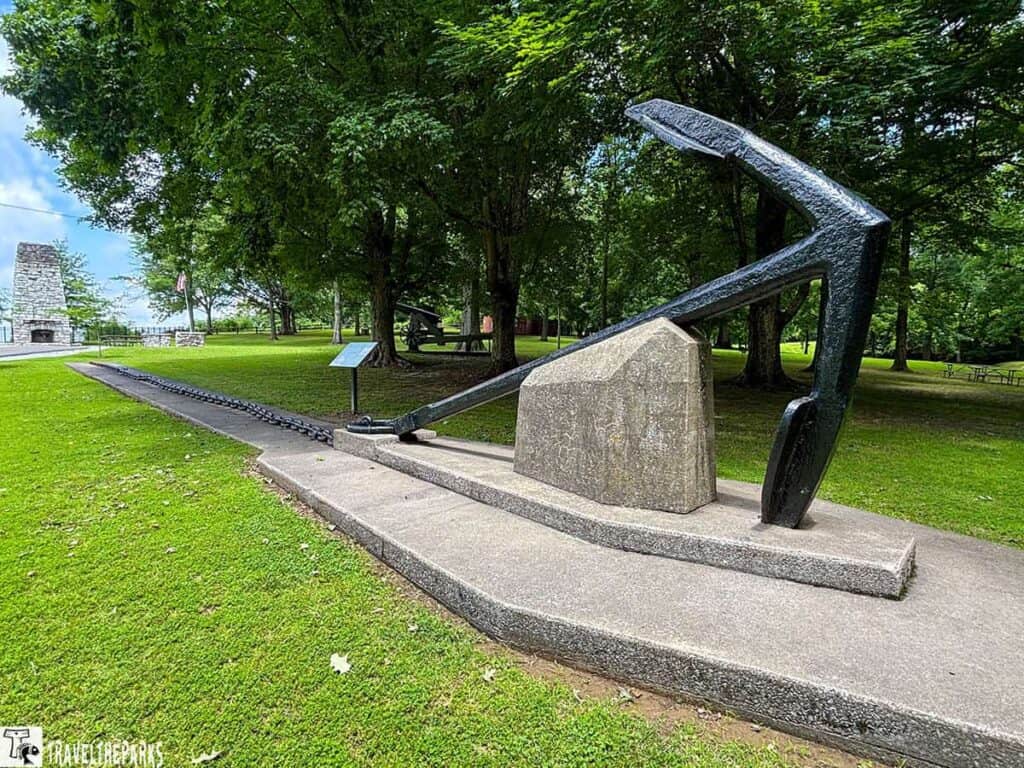 A large black anchor on a stone base, surrounded by green trees and a paved path in Belmont-Clermont State Park.