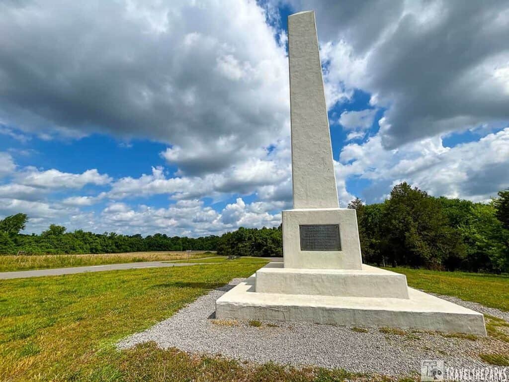 A tall white obelisk Artillary monument at Stones River National Battlefield, standing on a grassy area with trees in the background under a partly cloudy sky.

