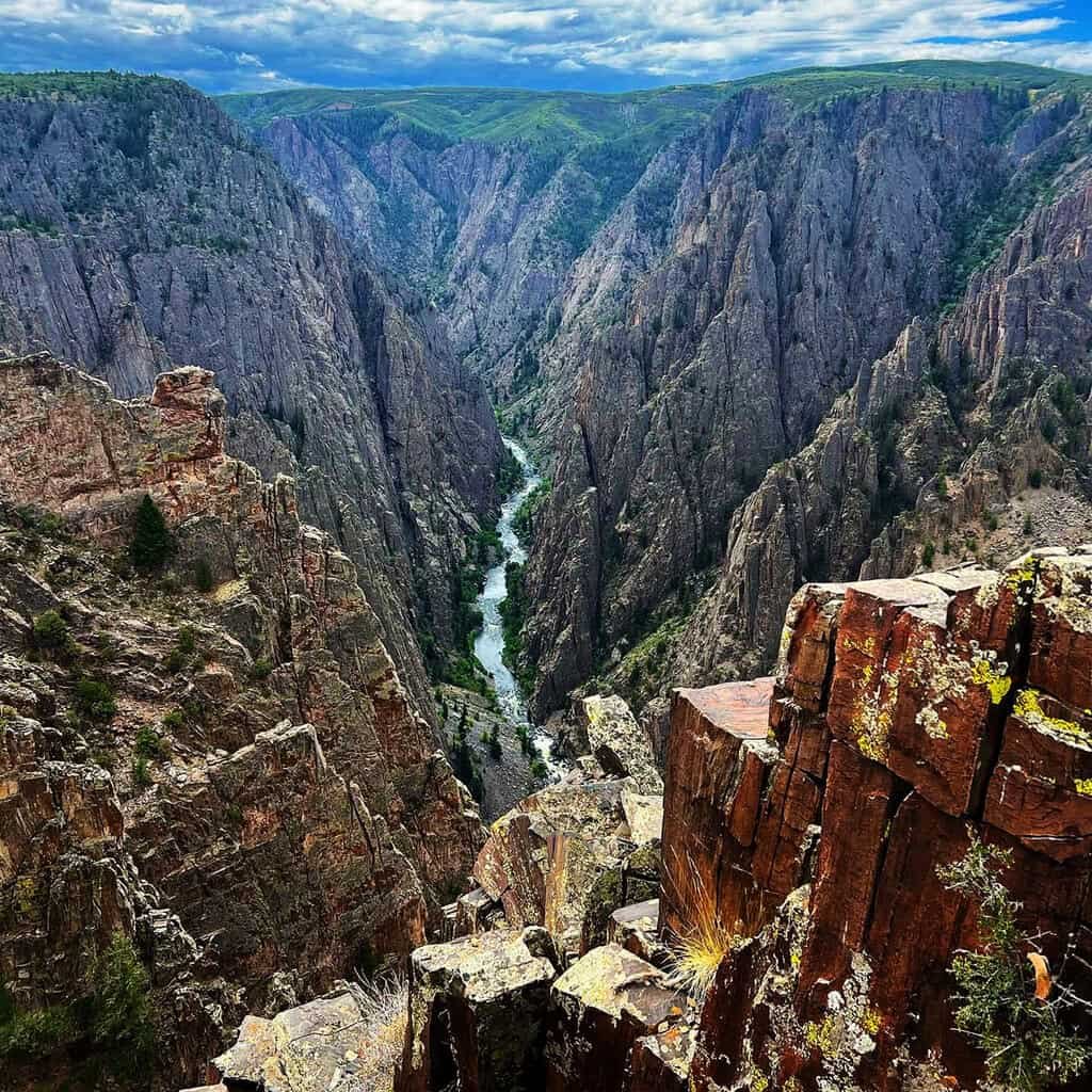 View of a deep, rugged canyon with steep rocky cliffs and a river flowing at the bottom. The sky is partly cloudy, and vegetation is sparse along the canyon edges.