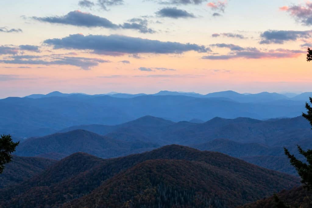 A panoramic view of blue-hued mountainous terrain at sunset, featuring rolling hills with autumn foliage in warm tones, under a sky of soft pink and blue.
