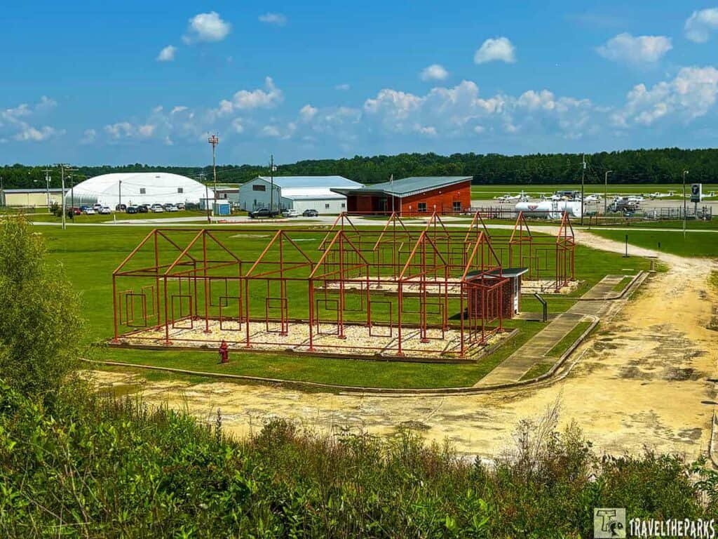 The Tuskegee Airmen National Historic Site, featuring red metal frames in the foreground and various buildings, including a large dome and aircraft, in the background against a blue sky.

