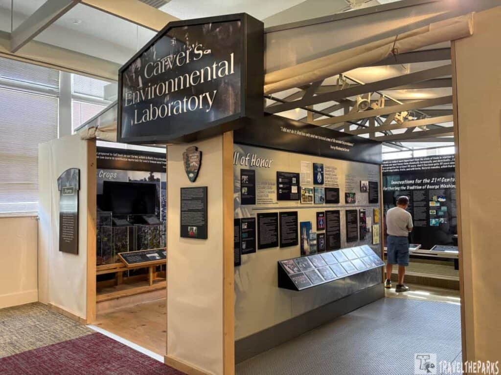 Interior view of Carver's Environmental Laboratory at Tuskegee Institute, featuring exhibits on agricultural innovations with a visitor interacting with the displays and a prominent sign overhead.

