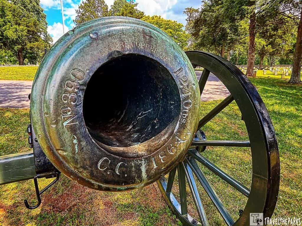 A close-up view of an antique cannon barrel, showing detailed engravings and surrounded by green grass and trees.

