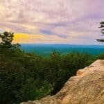 A rocky overlook surrounded by green trees, with rolling hills in the background under a pastel-colored sky at sunset.