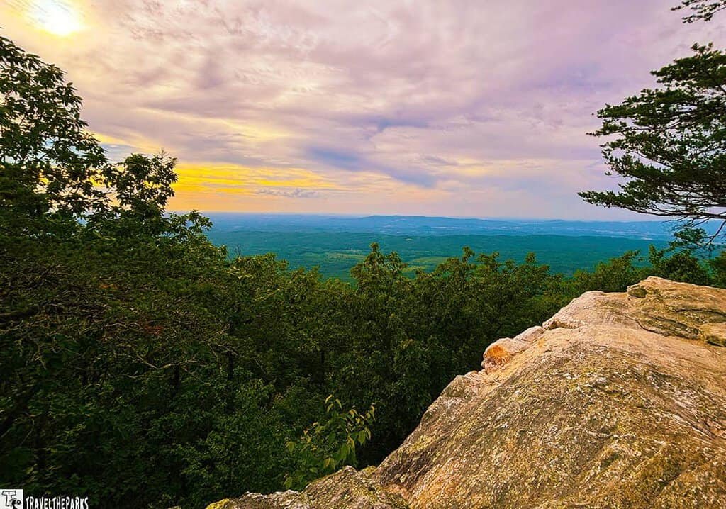 A rocky overlook surrounded by green trees, with rolling hills in the background under a pastel-colored sky at sunset.
