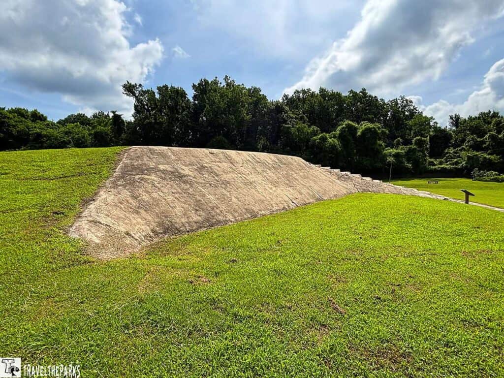 Chucalissa Mound is a large earthen structure with a sloped surface, surrounded by lush green grass and trees under a partly cloudy sky.