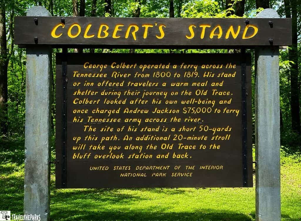 A wooden sign titled "COLBERT'S STAND" detailing the history of George Colbert's ferry service across the Tennessee River, surrounded by greenery.