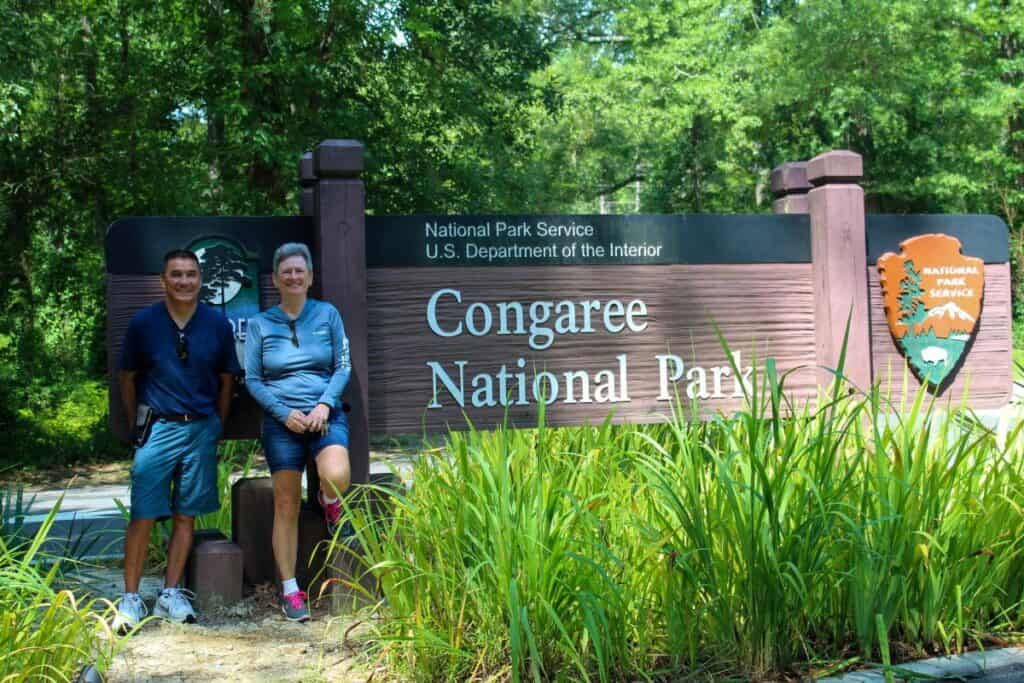 Two people stand smiling in front of the Congaree National Park sign, surrounded by lush greenery, under a clear, sunny sky.