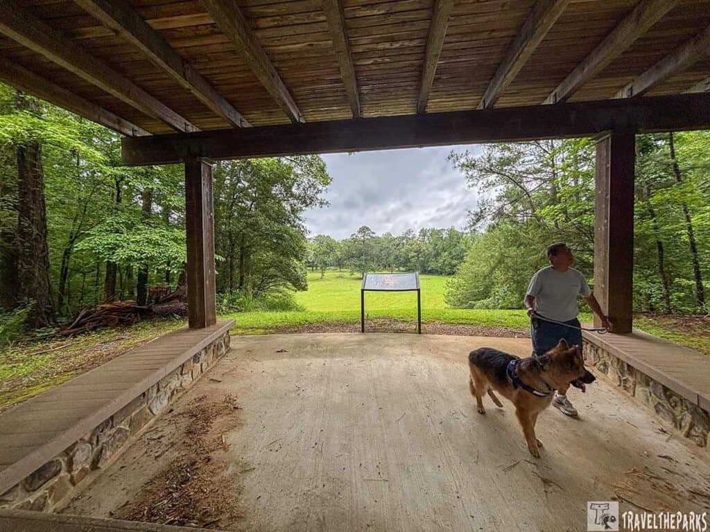 A covered pavilion with a person walking a German Shepherd dog, overlooking a green field and trees at Horseshoe Bend National Military Park.