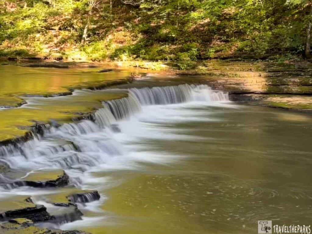 A waterfall cascades over rocky ledges into a calm pool, surrounded by green foliage and trees at David Crockett State Park.