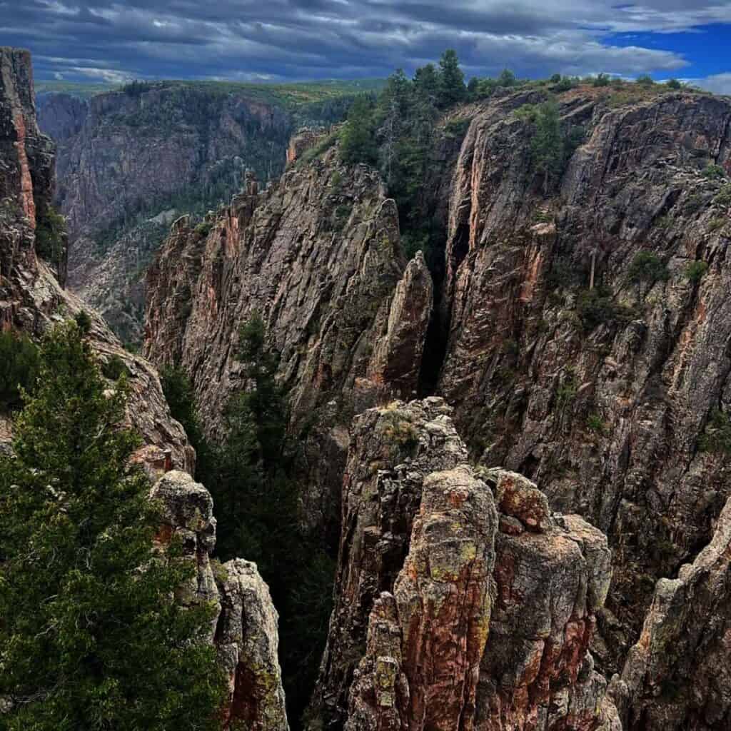 A dramatic landscape featuring a rugged canyon with steep, rocky cliffs. The scene is lush with greenery, as trees and shrubs cling to the rocky surfaces. Overhead, a cloudy sky creates a stark contrast with the vibrant natural elements below—perfect for a picturesque Colorado road trip to the Black Canyon of the Gunnison.