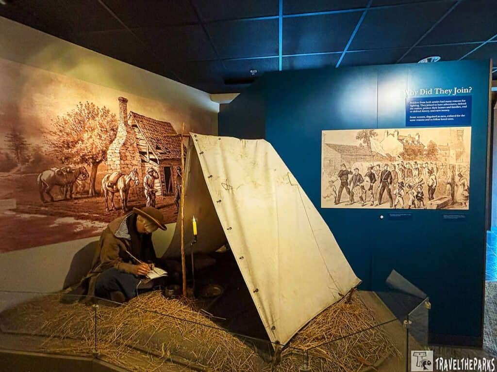 Diorama of Civil War camp life showing a soldier writing in front of a tent, with a mural of a cabin and horses in the background and an informational panel about soldiers' motivations.

