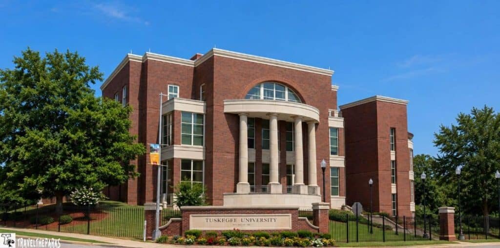 Exterior view of Tuskegee University building with red brick façade, columns at the entrance, surrounded by green lawns and trees under a clear blue sky.

