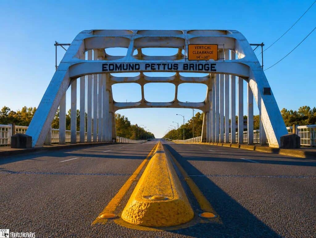 The Edmund Pettus Bridge, a large white arched structure, spans an empty roadway under a clear blue sky, with the name displayed prominently.