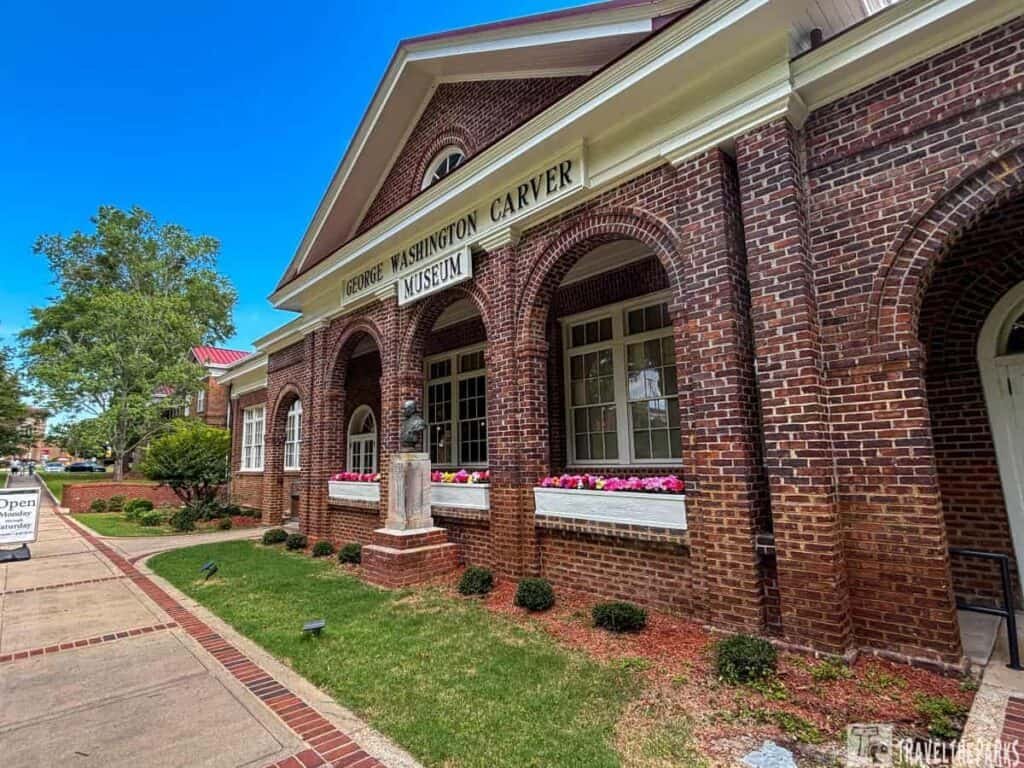 Exterior view of the George Washington Carver Museum, featuring brick architecture, large windows, a flower box, and a statue of Carver on a pedestal.


