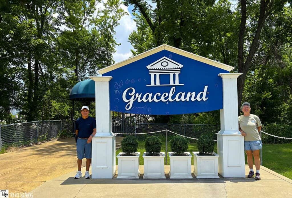 A blue and white "Graceland" sign with two people standing beside it, surrounded by greenery and planters.