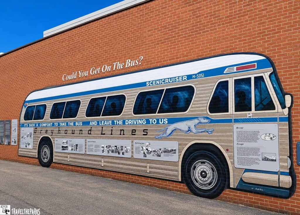 Mural of a vintage Greyhound bus on a brick wall, featuring silhouettes inside and text above reading, "Could You Get On The Bus?" At the bottom, informational plaques about Greyhound Lines are displayed.