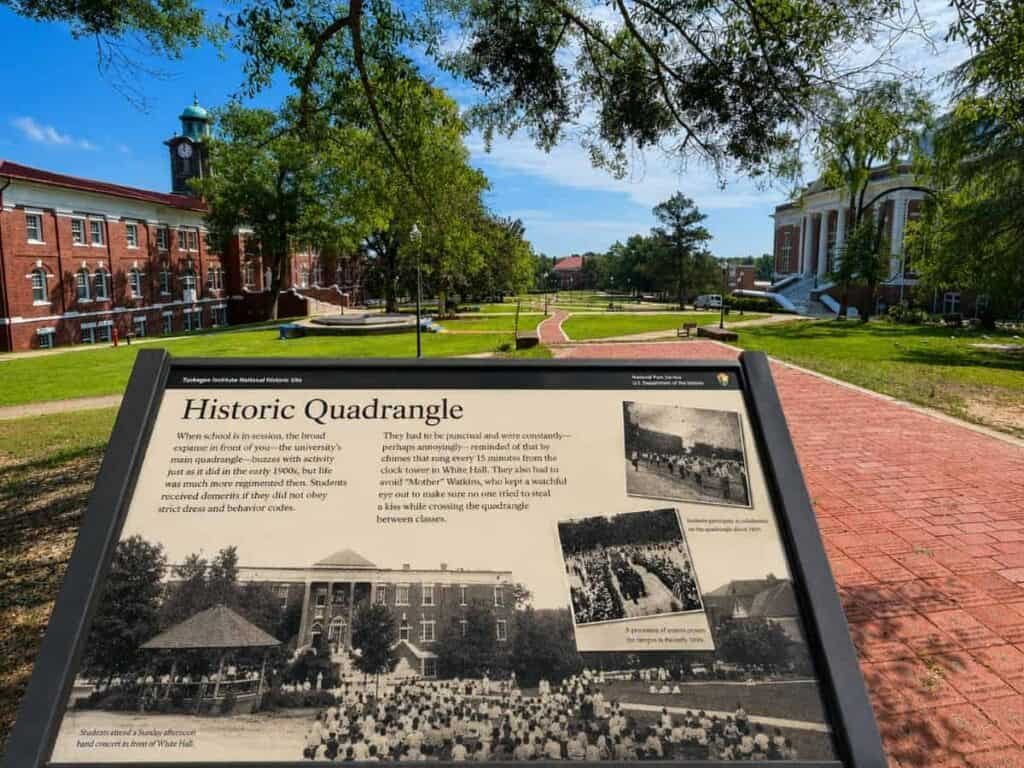 Historic Quadrangle at Tuskegee Institute featuring a sign with text and historical images, surrounded by green grass, pathways, and buildings under a blue sky.