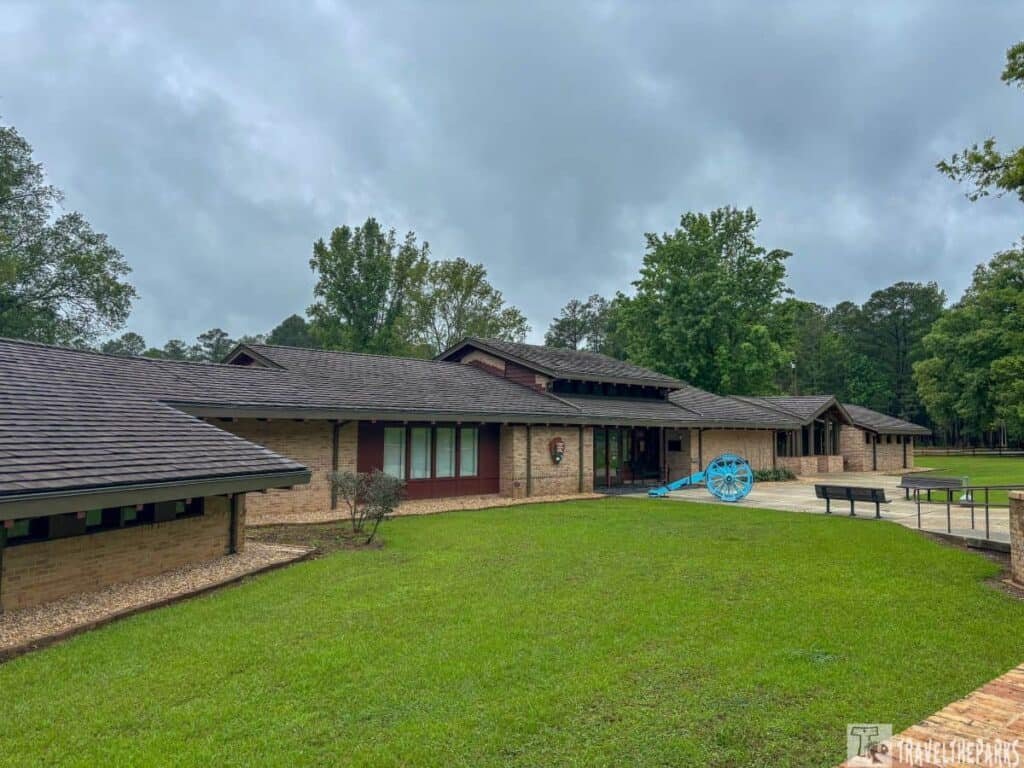 Visitor center at Horseshoe Bend National Military Park, featuring a brick building with a gabled roof, green lawn, blue cannon, and overcast sky.