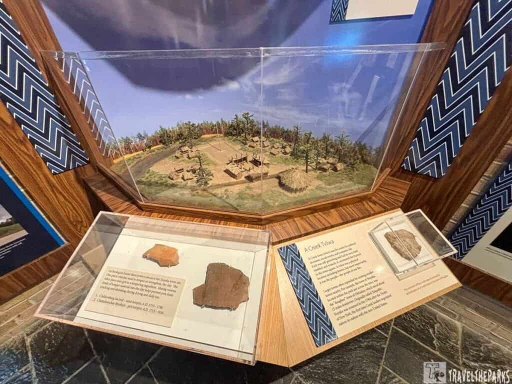 A three-dimensional model of historic structures and a natural landscape displayed in a glass case, accompanied by two information panels featuring pottery artifacts at Horseshoe Bend National Military Park.