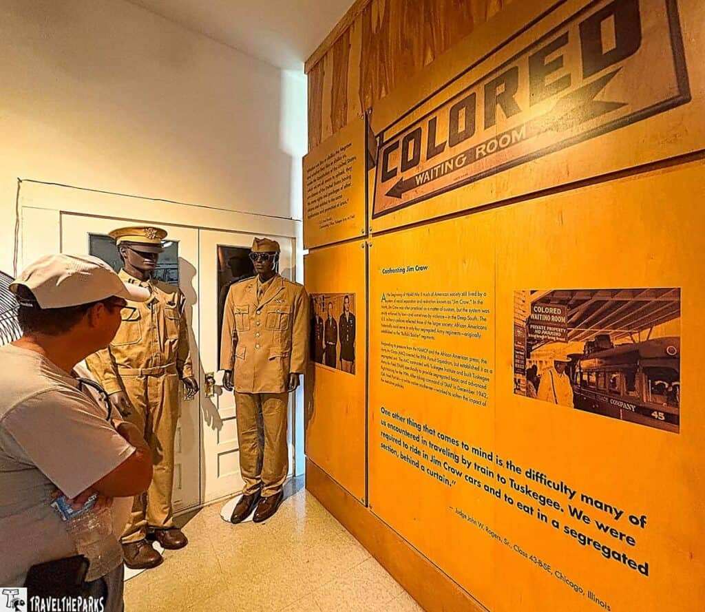 A museum exhibit featuring two mannequins in military uniforms and a wall sign reading "COLORED WAITING ROOM," with text discussing racial segregation in waiting areas. A viewer is looking at the display.

