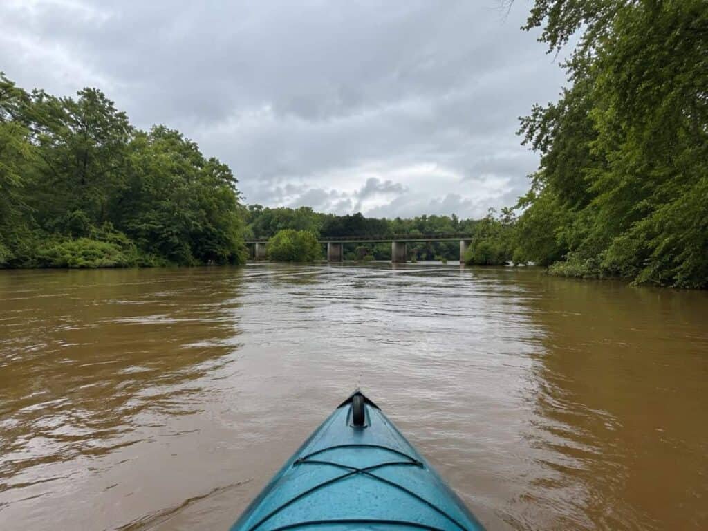 A turquoise kayak floats on the brown waters of the Tallapoosa River, with dense green trees on the left and a concrete bridge in the midground, all under an overcast sky.