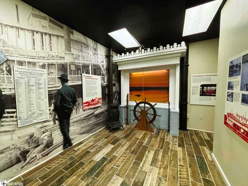 Interior of a riverboat exhibit with historical images, informational panels, and a ship's wheel by a sunset river scene.
Transcribed Text:
Table of Distances Chattahoochee, Apalachicola, Flint, and Chipola Rivers.
All Aboard the Steamboat Era.
A Riverboat's Pilothouse.
