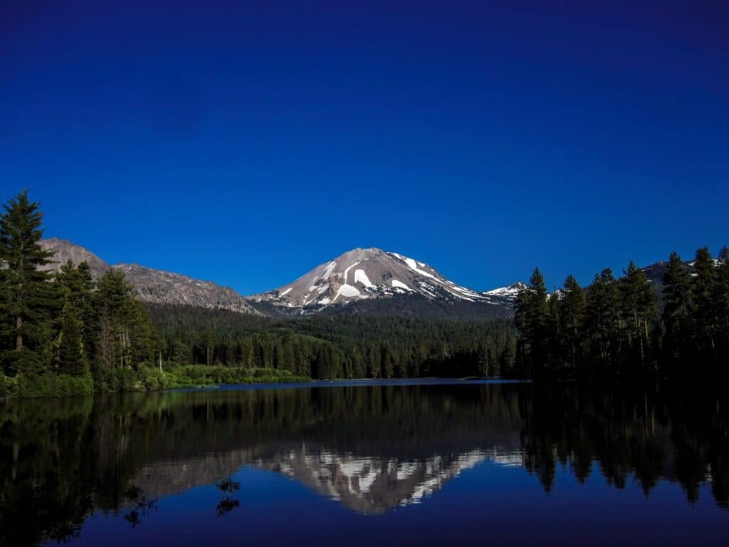 A serene lake reflecting Lassen Peak, a snow-capped volcano, under a clear blue sky, framed by dense evergreen trees.

