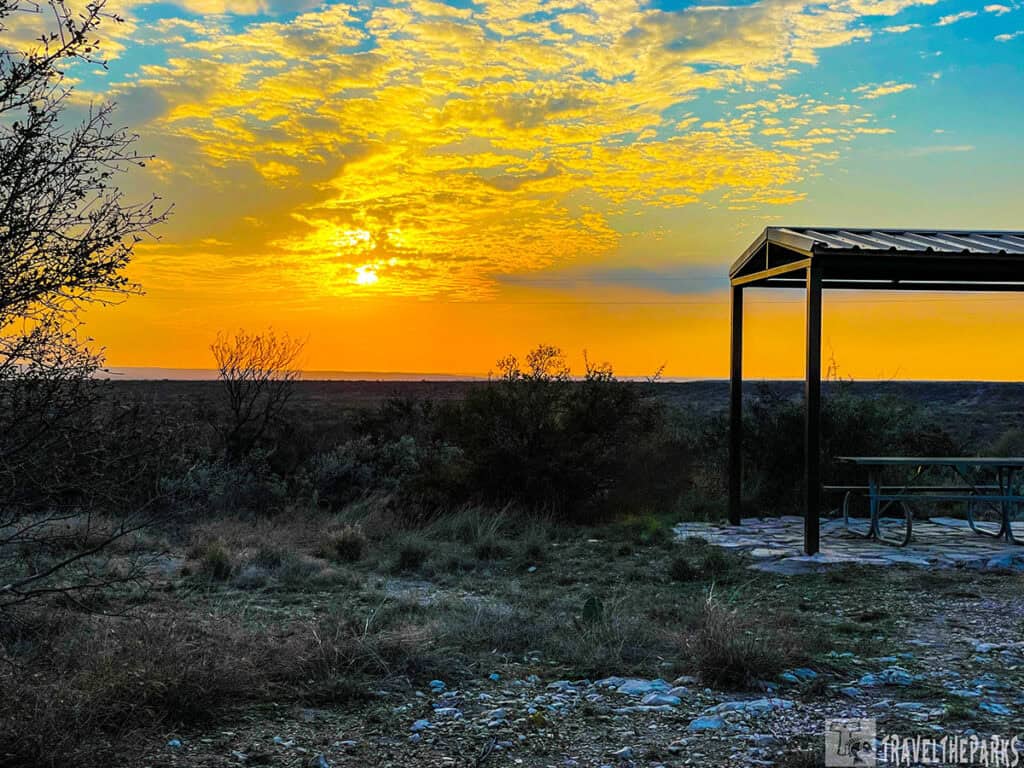 Sunset over a landscape featuring a metal shelter and picnic table, with dry grasses and shrubs in the foreground.