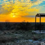 Sunset over a landscape featuring a metal shelter and picnic table, with dry grasses and shrubs in the foreground.