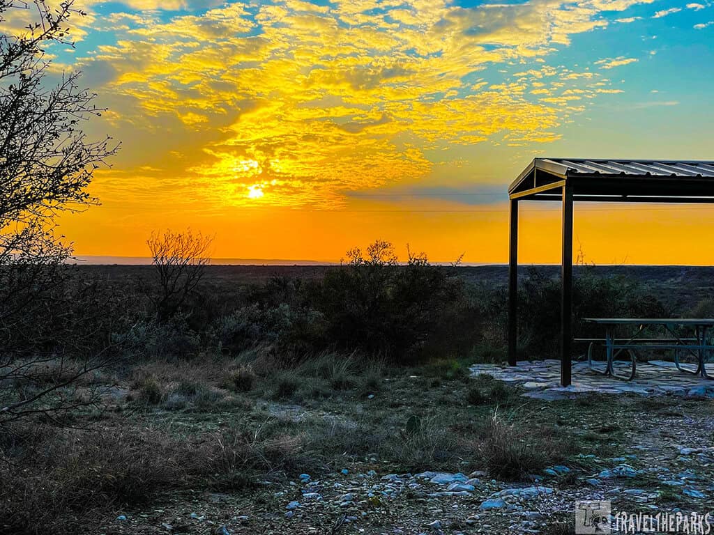 Sunset over a landscape featuring a metal shelter and picnic table, with dry grasses and shrubs in the foreground.