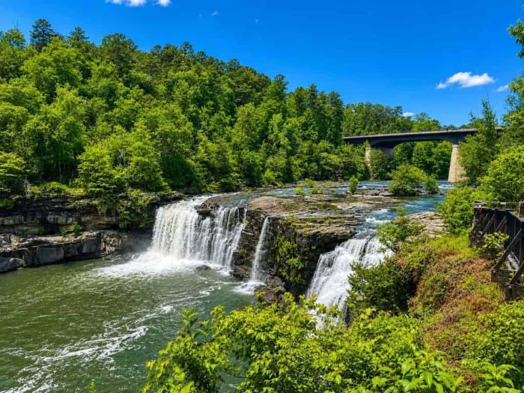 A waterfall cascades over rocky ledges into a river surrounded by lush green vegetation and a concrete bridge in the background under a clear blue sky.