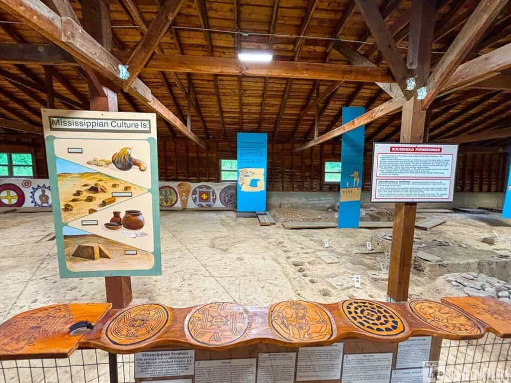 Interior view of Wickliffe Mounds museum displaying artifacts and informational panels about Mississippian culture, with wooden beams overhead and archaeological sites in the background.