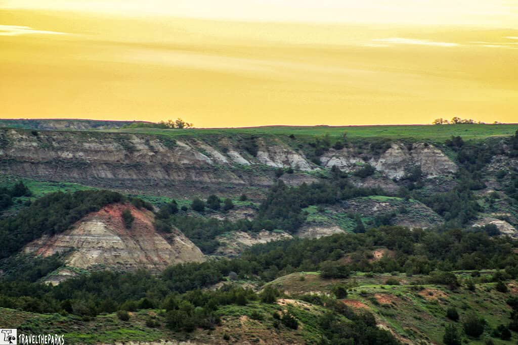 Panoramic view of Theodore Roosevelt National Park featuring rolling hills, layered rock formations, and a sunset sky with yellow and orange hues.

