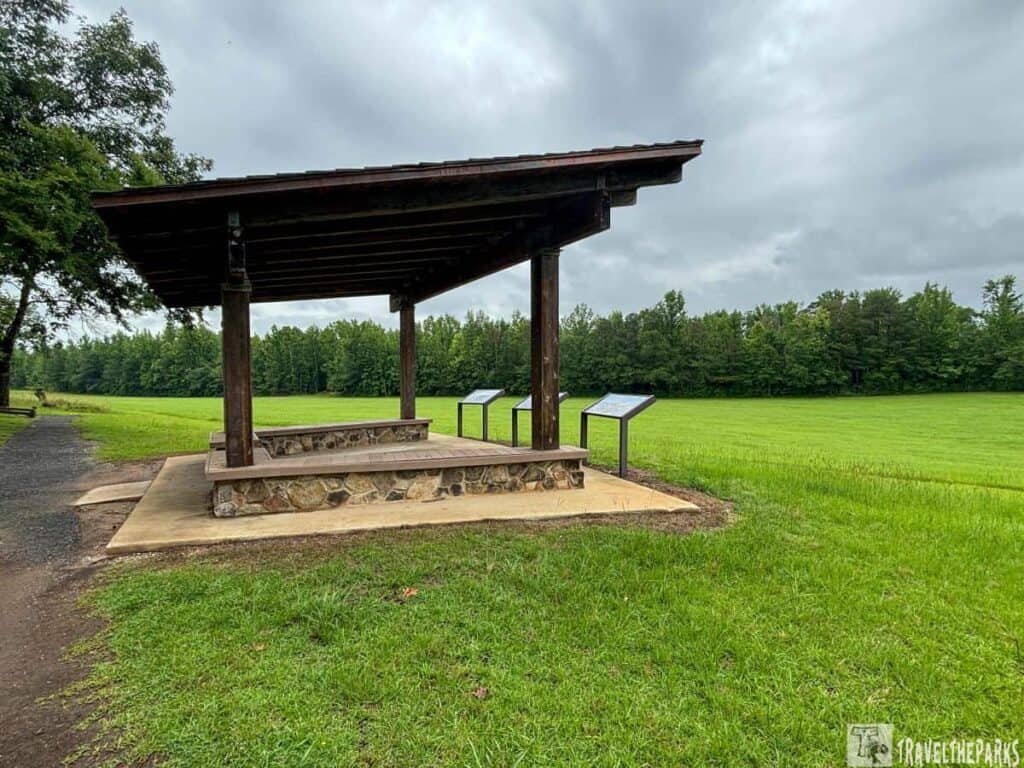 A wooden pavilion with a stone base at Horseshoe Bend National Military Park, surrounded by green grass and trees under a cloudy sky.
