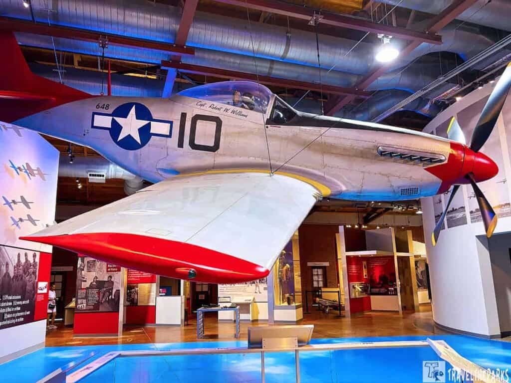 A P-51 Mustang plane, silver with a red tail and blue star insignia, is suspended in the Tuskegee Airmen National Historic Site. The surrounding museum features red and white display panels.

