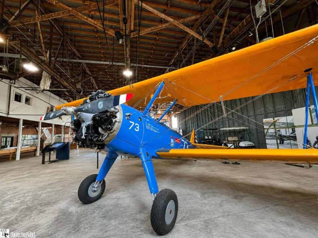 A vintage blue biplane with an orange upper wing displayed inside a hangar, showcasing its propeller and engine in a historical aviation setting.