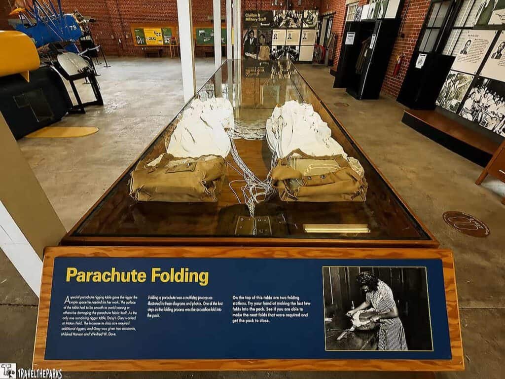 Exhibit at the Tuskegee Airmen National Historic Site featuring a glass table displaying two folded parachutes and their bags, with museum exhibits and a vintage aircraft in the background.

