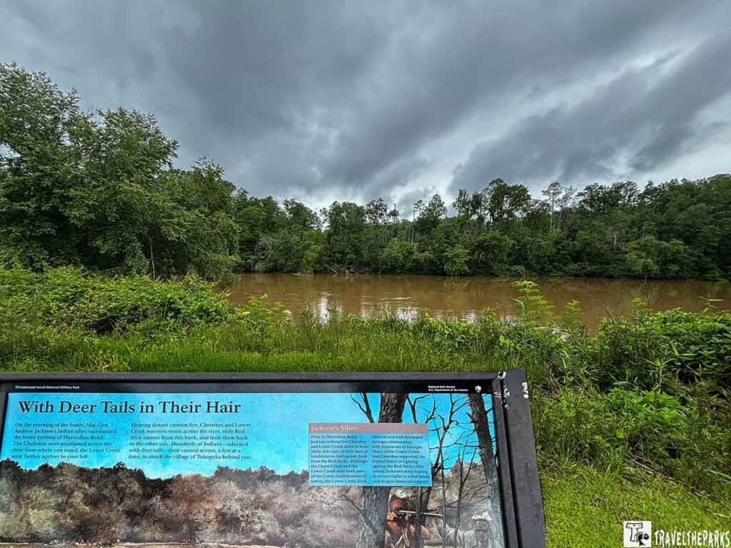 A view of Horseshoe Bend National Military Park with a prominent informational sign in the foreground, a muddy river, and dark, overcast clouds above.
Transcribed Text:
"With Deer Tails in Their Hair
On the morning of the battle, Maj. Gen. Andrew Jackson's Indian allies mentioned the former position of Hendrick's Beach. The Cherokees were positioned across the river from where you are standing; the Lower Creek were further upstream to your left."