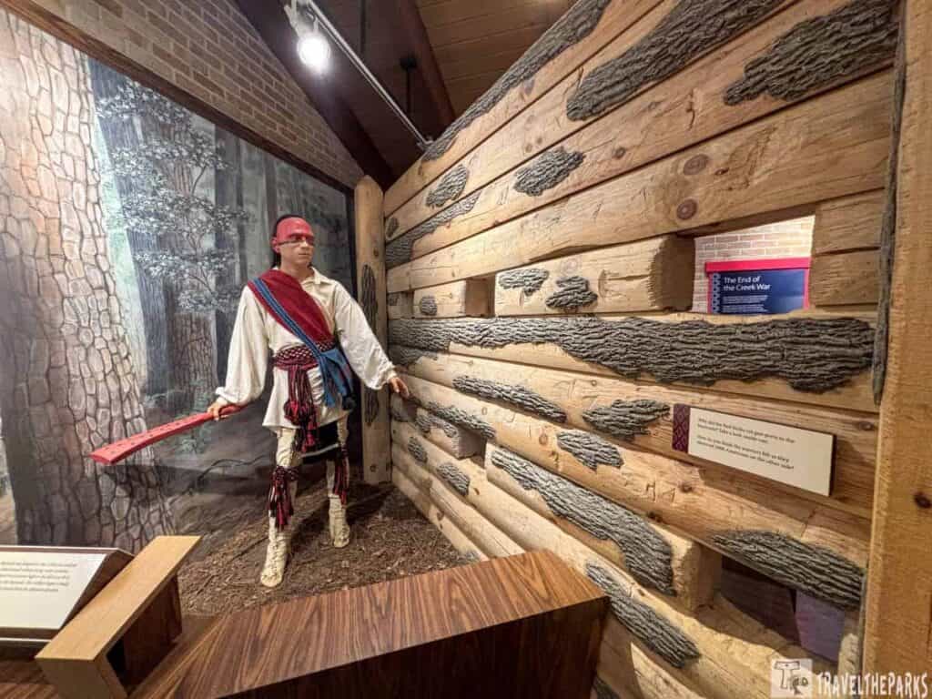 A life-size figure of a Red Sticks warrior dressed in traditional attire stands near a textured wooden wall in a historical exhibit at Horseshoe Bend National Military Park.