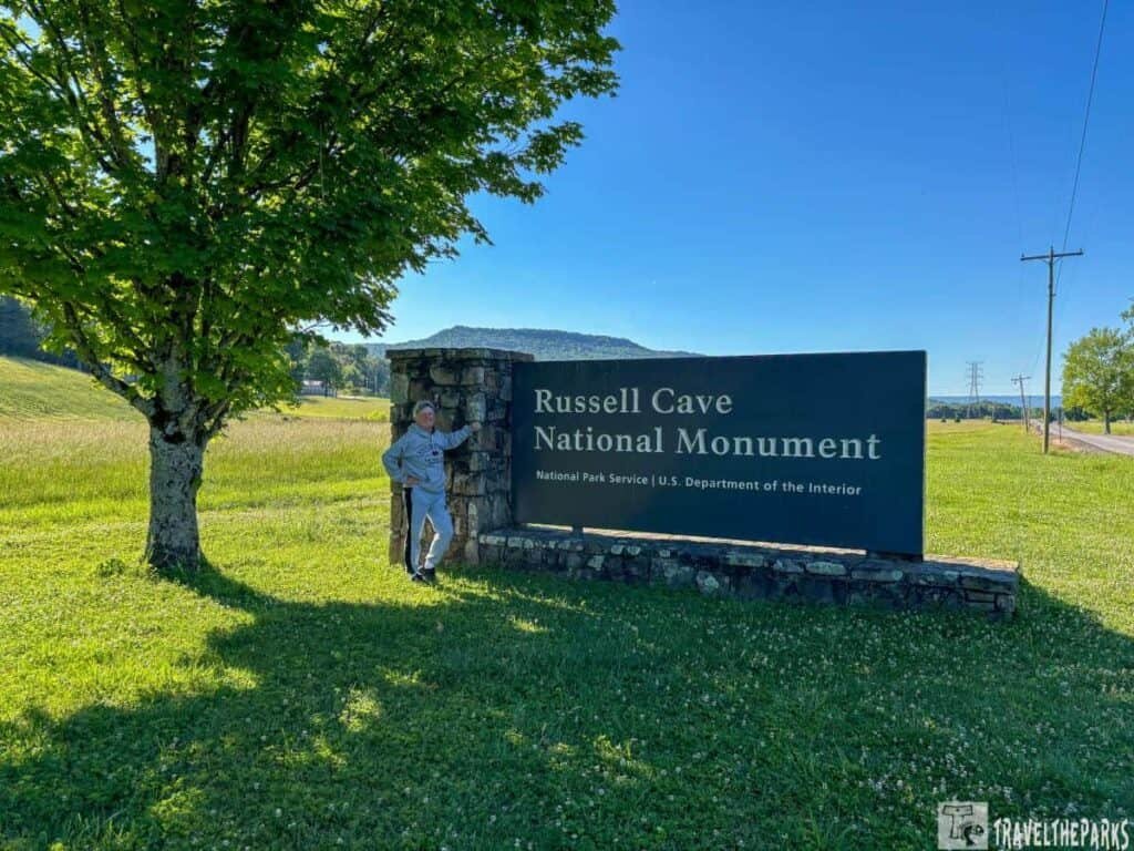 Person standing beside the sign for Russell Cave National Monument, with green grass and trees in the background under a clear blue sky.